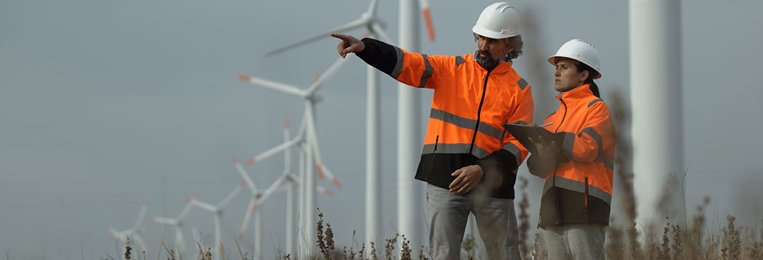 Two people wearing hard hats and orange jackets standing next to wind turbines.